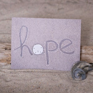 May include: A close-up of a card with the word "hope" written in the sand. A small white seashell is placed in the middle of the word.