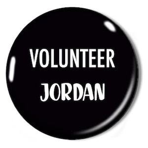 Könnte beinhalten: Ein schwarzer Knopf mit dem weißen Schriftzug "VOLUNTEER JORDAN". Der Knopf hat eine glänzende Oberfläche und eine leichte Reflexion. Der Text ist zentriert.