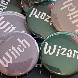 Może przedstawiać: Zbiór sześciu okrągłych, guzikowych znaczków z różnymi kolorami i wzorami. Znaczek przedstawiają słowa "Witch" i "Wizard" w stylu gotyckim.