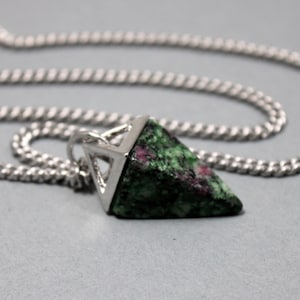 May include: A silver chain necklace with a triangular pendant. The pendant features a green and red stone set in a silver frame. The chain is a simple curb style, and the background is a solid gray.