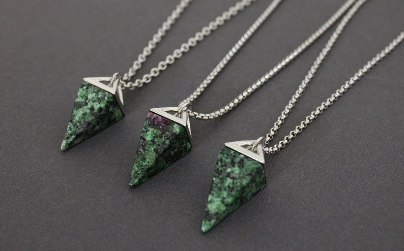 May include: Three necklaces with triangular pendants. Each pendant is made of green and red speckled stone, set in a silver-colored metal frame, and hangs from a delicate chain. The pendants have a geometric shape.