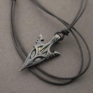 May include: A black leather cord necklace with a silver arrowhead pendant. The arrowhead has a detailed design with intricate lines and curves.