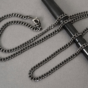 May include: A gunmetal-colored curb chain necklace with a lobster clasp. The chain is draped over a black pen on a gray surface. The necklace has a simple, classic design.
