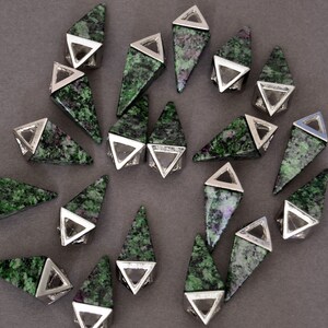 May include: A collection of gemstone pendants with a triangular shape. Each pendant features a green and red speckled stone set in a silver-colored metal frame. The pendants are arranged on a gray surface.