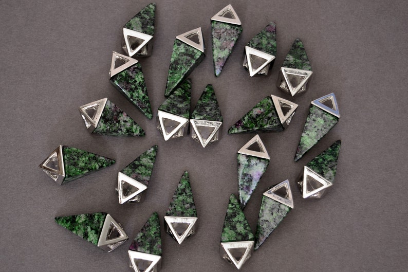 May include: A collection of gemstone pendants with a triangular shape. Each pendant features a green and red speckled stone set in a silver-colored metal frame. The pendants are arranged on a gray surface.