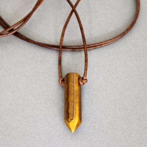 Peut inclure: Un collier pendentif avec un cristal cylindrique brun doré et une pointe. Le cristal est suspendu à un fin cordon en cuir marron. Le collier est présenté sur un fond gris neutre.