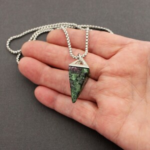 May include: A pendant necklace featuring a triangular, faceted gemstone with a mix of green and red hues. The gemstone is set in a silver-colored metal frame and hangs from a delicate, silver-toned chain. The necklace is displayed against a neutral background.