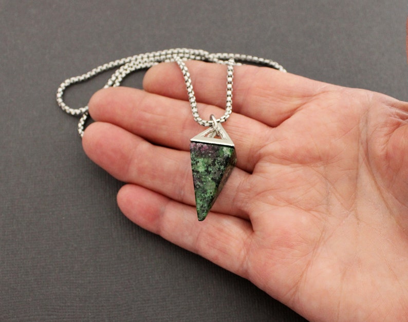 May include: A pendant necklace featuring a triangular, faceted gemstone with a mix of green and red hues. The gemstone is set in a silver-colored metal frame and hangs from a delicate, silver-toned chain. The necklace is displayed against a neutral background.