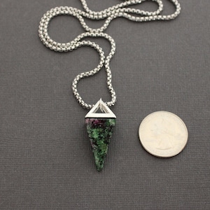 May include: A silver chain necklace with a pendant featuring a triangular silver frame and a green and red stone. A quarter is next to the pendant for size comparison. The necklace is on a dark gray surface.
