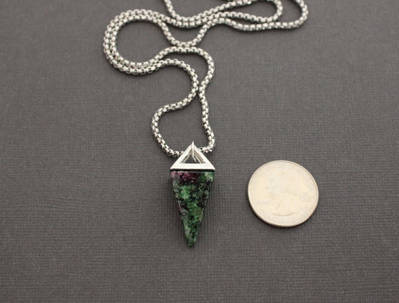 May include: A silver chain necklace with a pendant featuring a triangular silver frame and a green and red stone. A quarter is next to the pendant for size comparison. The necklace is on a dark gray surface.