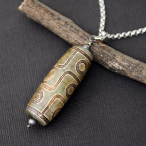 Peut inclure: Un pendentif cylindrique en pierre vert clair et beige orné de motifs circulaires et linéaires gravés. Le pendentif est suspendu à une chaîne en métal argenté et est surmonté de petites perles métalliques aux extrémités supérieure et inférieure.