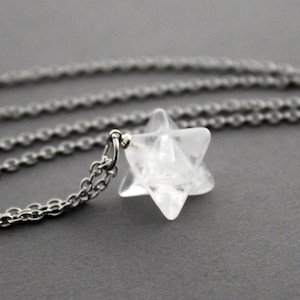 May include: A clear, faceted, star-shaped crystal pendant suspended from a silver-toned metal chain. The pendant features a geometric, multi-pointed design with smooth, translucent surfaces that catch the light, attached to the chain by a small metal ring.