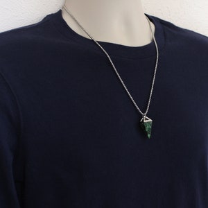 May include: A silver chain necklace with a geometric pendant. The pendant is a dark green stone with a silver metal frame. The person in the image is wearing a navy blue long-sleeve shirt.