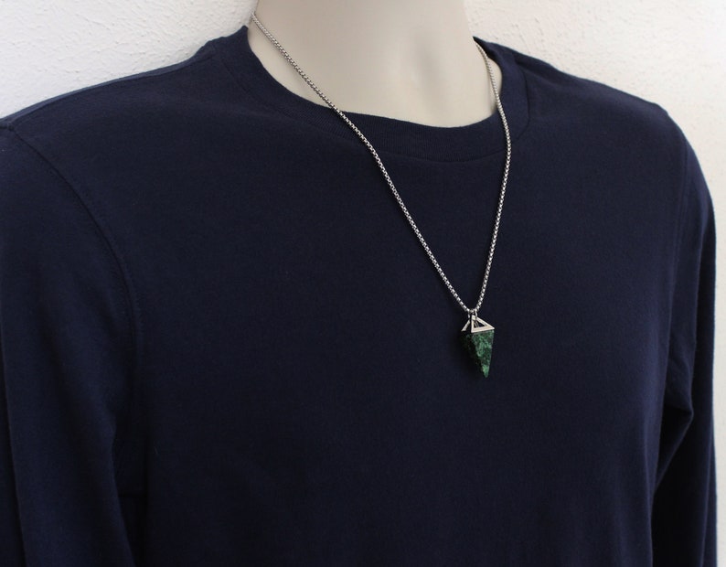 May include: A silver chain necklace with a geometric pendant. The pendant is a dark green stone with a silver metal frame. The person in the image is wearing a navy blue long-sleeve shirt.