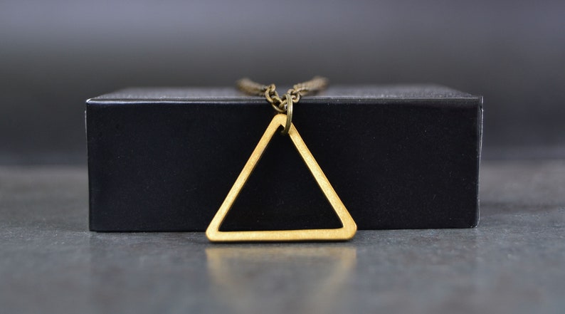 Peut inclure: Un collier avec un pendentif en forme de triangle dor&eacute; sur une bo&icirc;te noire. Le triangle est creux et a un design simple.