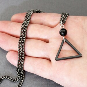 May include: A black chain necklace with a black triangle pendant and a black bead. The necklace is on a hand.