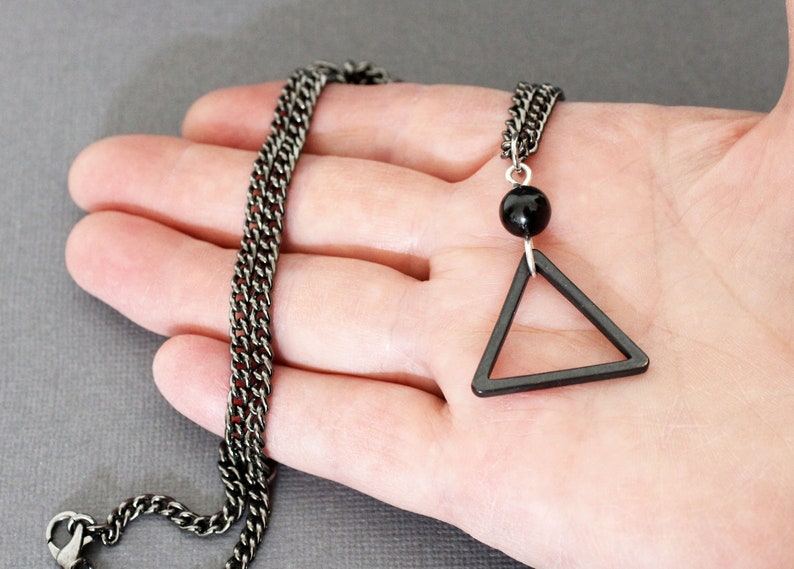 May include: A black chain necklace with a black triangle pendant and a black bead. The necklace is on a hand.