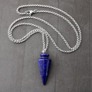 May include: A silver-colored chain necklace with a blue lapis lazuli pendulum pendant. The pendant is a deep blue color with a pointed shape. The chain is made of small, round links and has a clasp.