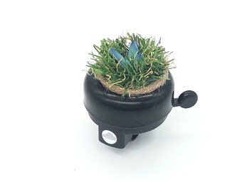 Peut inclure: Une sonnette de vélo noire avec une petite plante en pot sur le dessus. La plante a de l'herbe verte et des fleurs bleues.