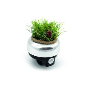 May include: A silver bicycle bell with a black base, topped with a miniature garden of green grass and a small red accent. The bell has a natural rope border around the grass.