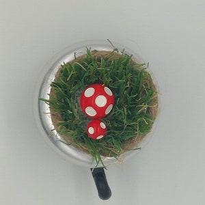 May include: A whimsical, miniature terrarium featuring two red and white spotted mushrooms nestled in green grass. The terrarium is set in a silver-colored, circular container with a black handle.