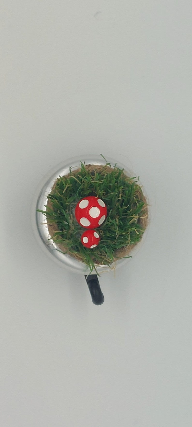 May include: A whimsical, miniature terrarium featuring two red and white spotted mushrooms nestled in green grass. The terrarium is set in a silver-colored, circular container with a black handle.