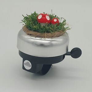 May include: Silver bicycle bell with a black lever and mounting bracket. The top of the bell features a small patch of green artificial grass with two red mushroom decorations with white spots. A charming accessory.