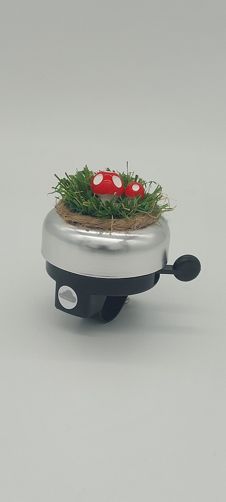May include: Silver bicycle bell with a black lever and mounting bracket. The top of the bell features a small patch of green artificial grass with two red mushroom decorations with white spots. A charming accessory.