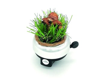 Op de afbeelding: Een zilveren fietsbel met een bruin luiaardfiguur genesteld in groen gras. De bel heeft een zwarte hendel en een klein, wit wolkontwerp op de basis. Het gras zit in een kleine, bruin gevoerde pot.
