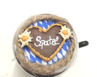 Op de afbeelding: Een zilveren fietsbel met een bruine hartvormige decoratie. Het hart heeft een witte inscriptie met de tekst "Spaetzle" en is omgeven door een blauw-wit geruit patroon. De bel is versierd met een bruin touw.
