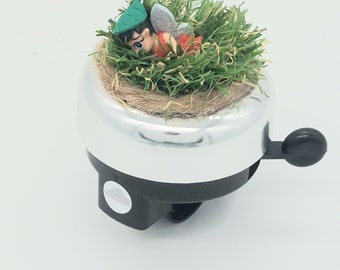 Peut inclure: Une sonnette de vélo argentée avec une poignée noire. La sonnette est décorée d'une petite figurine de fée assise dans un lit d'herbe verte.