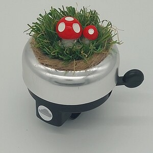 May include: Silver bicycle bell with a black lever and base. The bell is decorated with a bed of green faux grass and two red mushrooms with white spots. A charming, whimsical design.