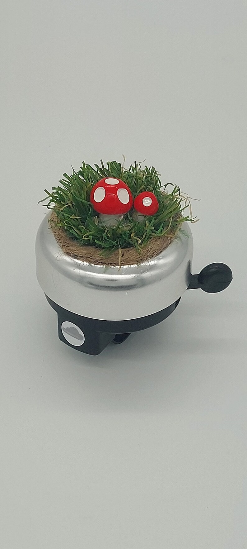 May include: Silver bicycle bell with a black lever and base. The bell is decorated with a bed of green faux grass and two red mushrooms with white spots. A charming, whimsical design.