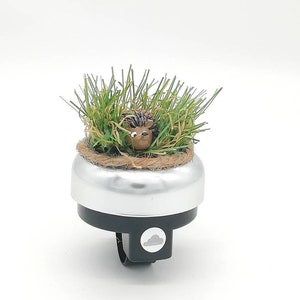 Könnte beinhalten: Eine silberne Fahrradklingel mit schwarzem Sockel und einer kleinen Igel-Figur in künstlichem grünen Gras. Der Igel hat ein hellbraunes Gesicht und dunkelbraune Stacheln.