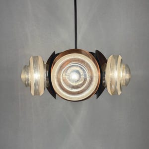 Może przedstawiać: Nowoczesna lampa wisząca z centralnym dyskiem w kolorze miedzi, otoczonym czarnymi metalowymi akcentami i przezroczystymi szklanymi elementami. Oprawa ma unikalny design z trzema źr&oacute;dłami światła, tworząc ciepłą, zachęcającą poświatę.