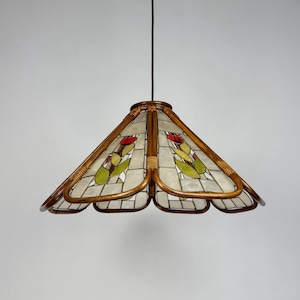 Op de afbeelding: Hanglamp met een gebrandschilderde glazen kap met rode en groene bloemmotieven. De kap is omlijst met een geweven rotan materiaal en de lamp hangt aan een bruin snoer. De lamp heeft een unieke, vintage uitstraling.
