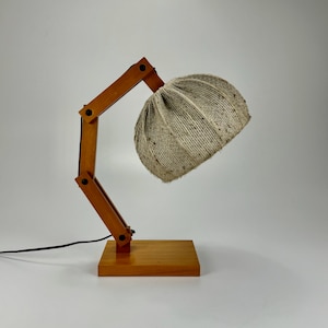 May include: A wooden desk lamp with a natural fiber shade. The lamp features a rectangular base and an adjustable arm with black joints. The shade is a light beige color and has a textured appearance. A black power cord extends from the base.