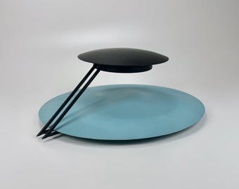 May include: A black and light blue table lamp with a circular base and a circular shade. The lamp has a unique design with two black metal arms extending from the base to the shade.