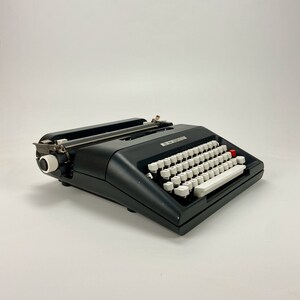 以下が含まれることがあります： 白いキーボードと赤いボタンが付いた黒いビンテージタイプライター。タイプライターは金属製で、クラシックなデザインです。
