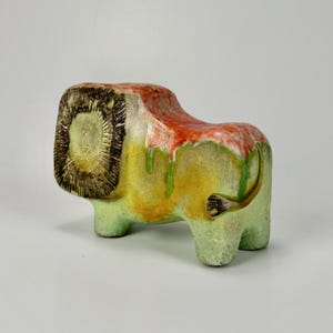 Puede incluir: Escultura de león de cerámica con melena y cuerpo texturizados. La cabeza del león es marrón, con una cara verde claro. El cuerpo hace la transición de amarillo a verde, con detalles en rojo y blanco. La cola es curva y la escultura tiene cuatro patas cortas.