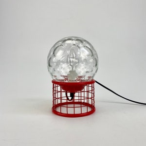 May include: A red metal table lamp with a clear glass globe shade. The globe has a textured surface and a white light bulb inside.