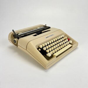 以下が含まれることがあります： 黒色のキャリッジリターンレバーと赤いキーが付いたベージュのOlivetti Lettera 36 タイプライター。タイプライターは、クリーム色のキーが付いた標準のQWERTYキーボードです。