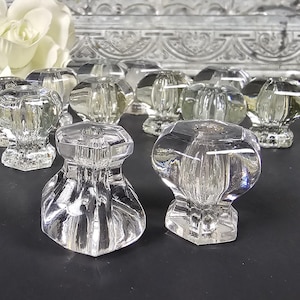 May include: Eight clear glass cabinet knobs with a faceted design. The knobs are arranged in a semi-circle on a black surface.