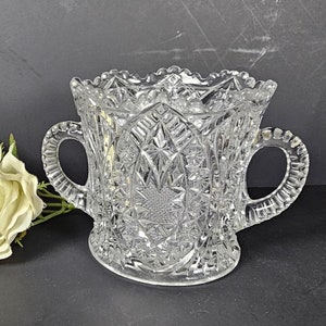 May include: A clear glass vase with two handles and a cut glass design. The vase is sitting on a black surface with a white rose.