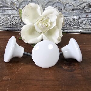 May include: Three white ceramic knobs with a round, flat top and a slightly raised edge. The knobs are on a wooden surface.