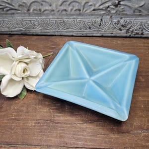 May include: A square, light blue ceramic dish with a geometric design. The dish has a raised, folded appearance and is displayed on a wooden surface. A white flower with green leaves is next to the dish.
