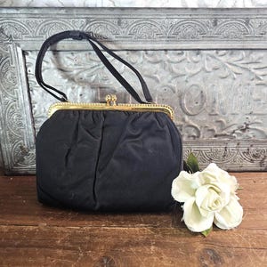 May include: A black fabric handbag featuring a gold-toned metal frame with a kiss-lock closure. The bag has a single black handle and a soft, gathered texture. A white flower is positioned next to the base of the handbag on a wooden surface.