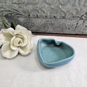 May include: A small, light blue ceramic ashtray with a unique, organic shape. A white rose is next to the ashtray. The ashtray has a smooth, matte finish and a small indentation on one side.