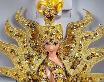 May include: A golden Barbie doll with a sun-inspired costume. The doll has blonde hair and is wearing a golden crown, a golden dress with sequins, and a golden cape with a sunburst design.