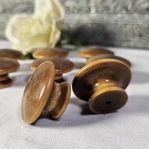 May include: A collection of wooden cabinet knobs with a mushroom-shaped design. The knobs are made of light brown wood with a darker brown stain on the top. The knobs are arranged on a white surface.